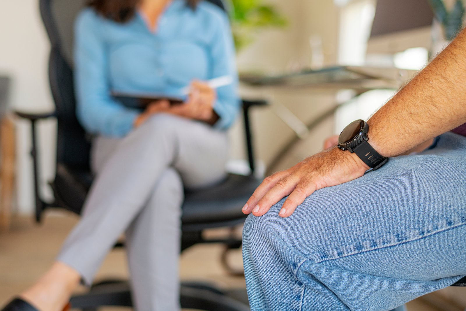 Therapeutin und Patient sitzen sich gegenüber. Die Therapeutin hält Stift und Notizblock.