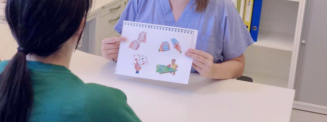 Logopädin sitzt an Tisch in freundlichem Büro, hält Schaubild in den Händen und erklärt einer Patientin etwas.