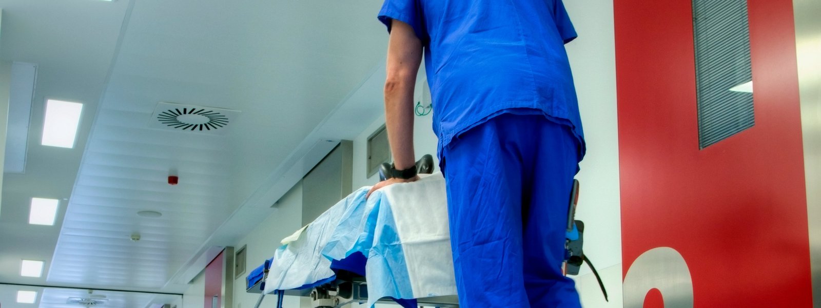Ein:e Mitarbeiter:in in blauer Arbeitskleidung schiebt ein Krankenbett in einem hell erleuchteten Gang.