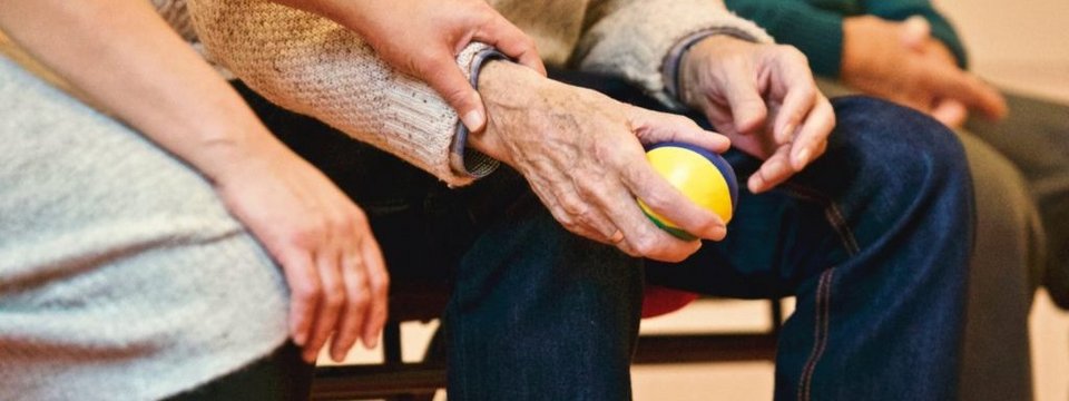Eine ältere Person hält einen bunten Ball und wird dabei von einer anderen Hand unterstützt.