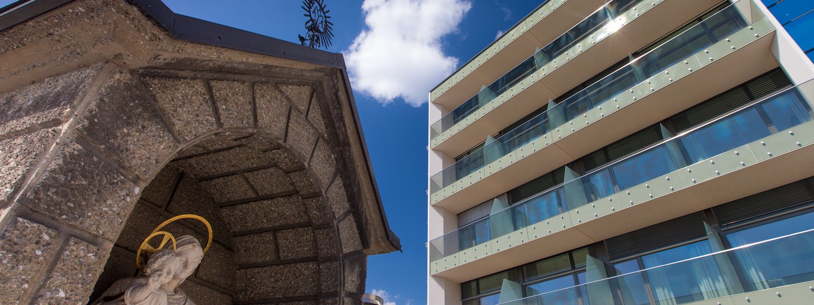 Kleine steinerne Kapelle vor modernem Krankenhausgebäude mit Glasbalkonen. Im Hintergrund strahlend blauer Himmel.