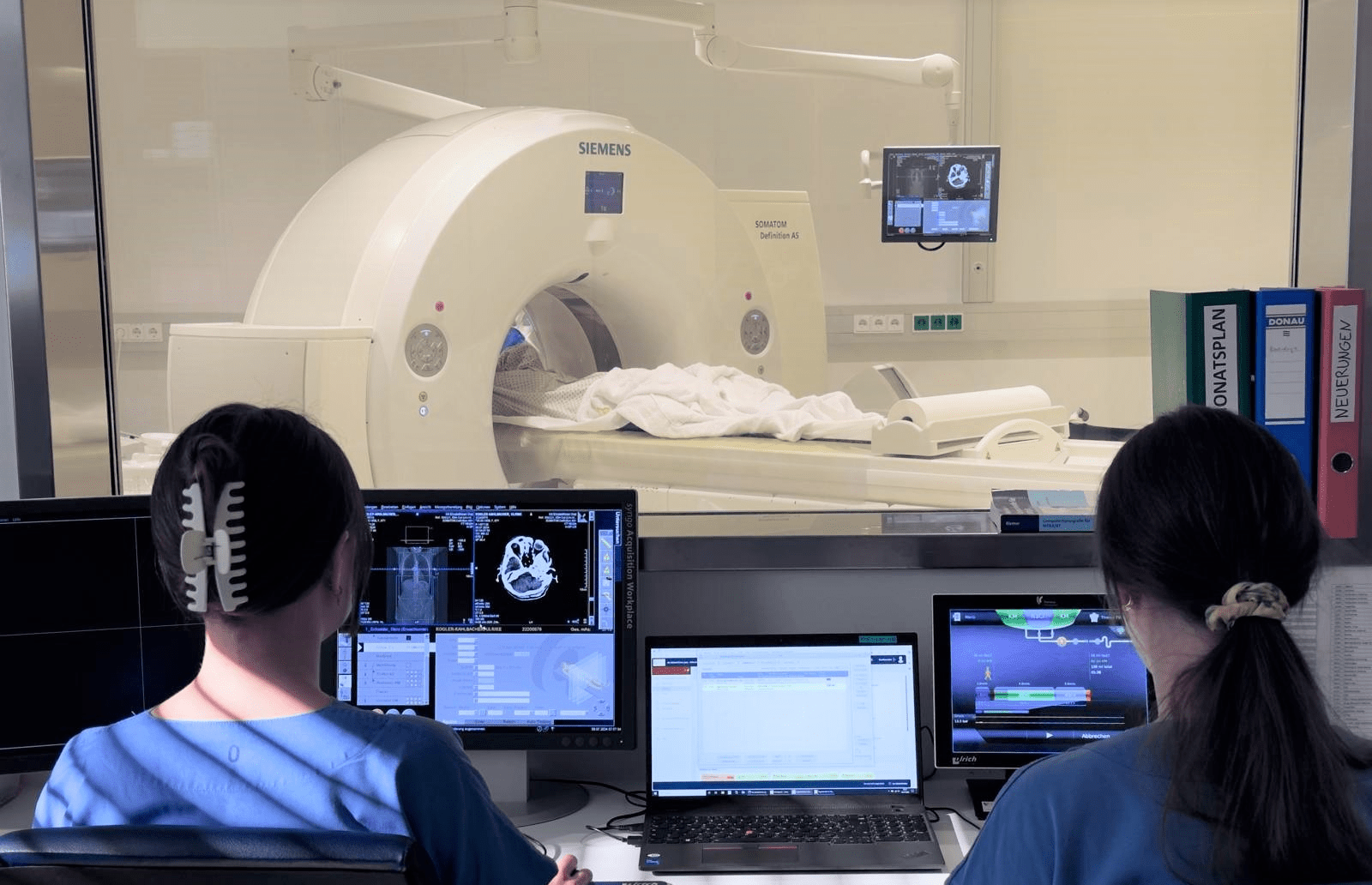 Zwei Radiologinnen vor den Bildschirmen, im Hintergrund das CT-Gerät.