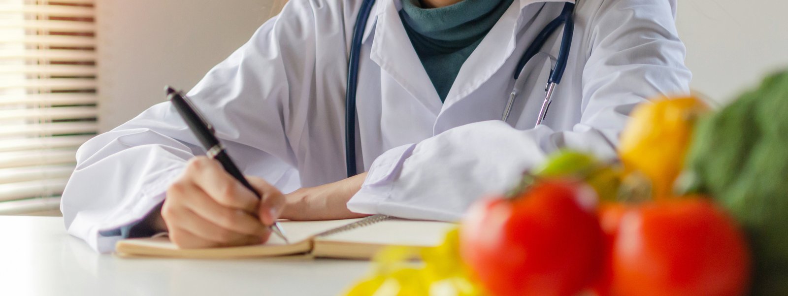 Diätolog:in sitzt im Büro, schreibt auf Block mit Gemüse im Vordergrund.